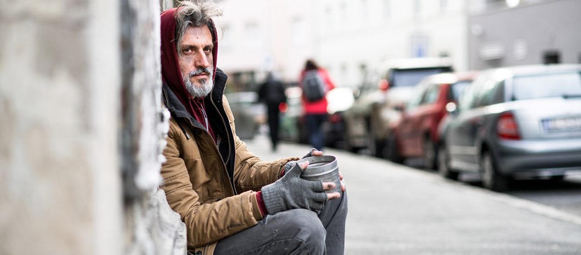 A side view of homeless beggar man sitting in front of wooden gate outdoors in city asking for money donation.